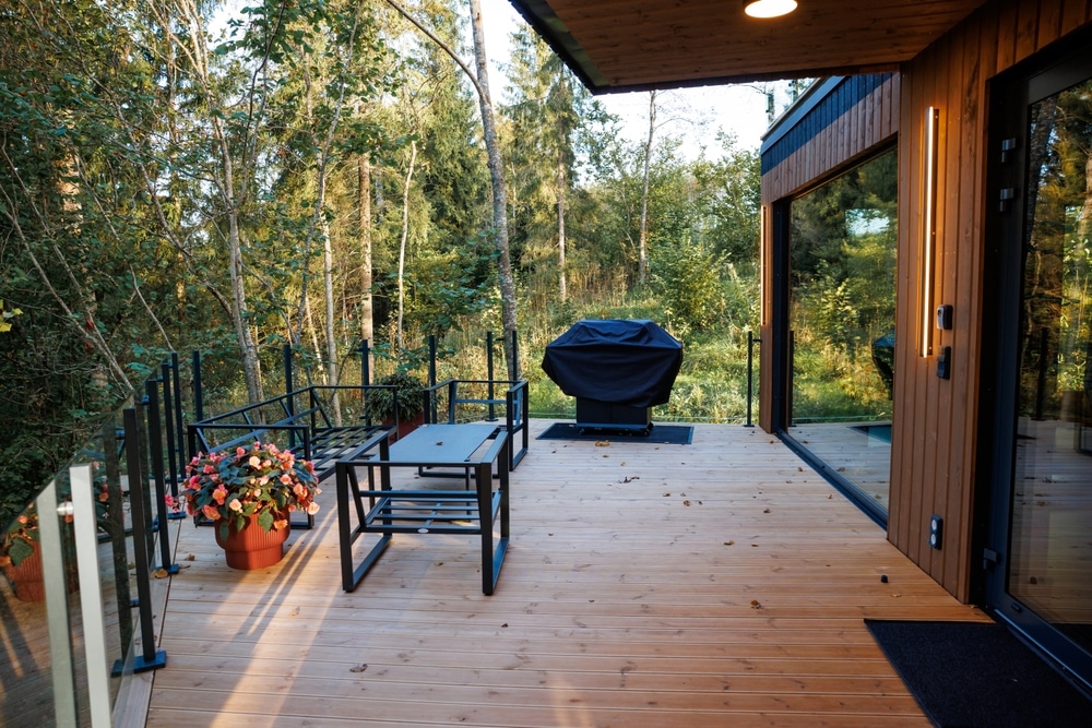 A modern wooden deck features black metal furniture, a grill, potted flowers, glass railings, and warm lighting, surrounded by a forested area.